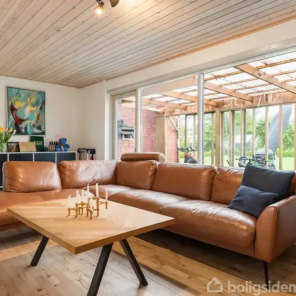 En brun hjørnesofa står i stuen, der er lyst indrettet med trægulve og loft. Foran sofaen er et træbord med lysestager, og store vinduer viser en terrasse og have.