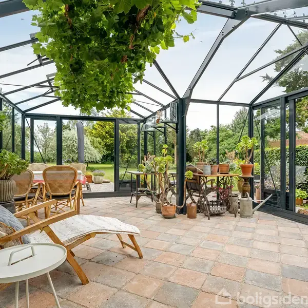 En udestue med glasvægge og loft fyldt med planter og møbler, som liggestole og småborde. Den omgivende have ses gennem glasset, med grønne træer og buske.