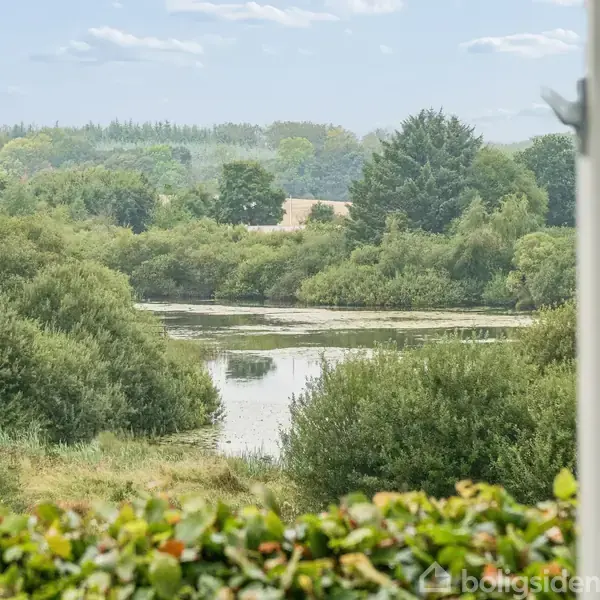 Et vindue med udsigt over en frodig dal med træer og en lille sø i midten. Landskabet strækker sig mod en skovrig horisont.