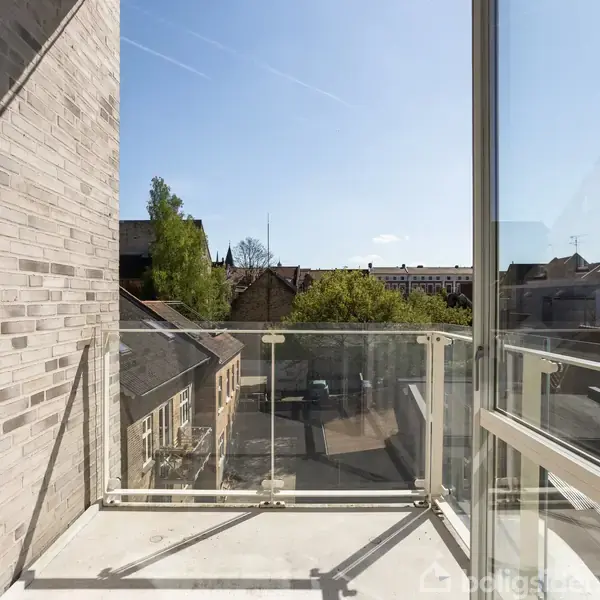 Balkon med glasrækværk, ingen møbler, vender mod byens hustage og en blå himmel. Sollys skinner ind på betongulvet.