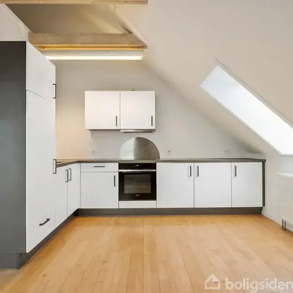 Køkken med hvide skabe og en ovn placeret under et skråt loft. Trægulv og stort skråvindue på højre side. Radiator under vinduet. Ingen tekst tilgængelig.