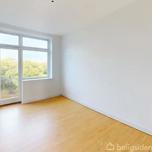 Et tomt værelse med lyst trægulv, en stor hvid væg og et vindue med dør udsyn til grønne trætoppe. Radiator monteret på væggen ved vinduet.