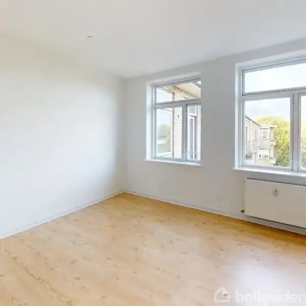 Et tomt værelse med trægulv og hvide vægge. To store vinduer slipper lys ind. Radiator under det ene vindue ses i en minimalistisk indretning i en lys lejlighed.