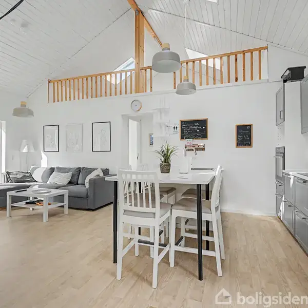 Stue med sofa og sofabord ved glasskydedør. Spisebord med stole i midten. Køkkenskabe langs højre væg. Lyse trægulve, højt til loftet med synlige bjælker, vægkunst og grønne planter.