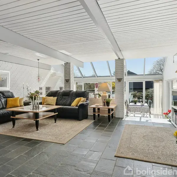 Stue med sorte lædersofaer og gule puder, stående på tæpper på mørkt stengulv. Store vinduer og glasdøre giver udsigt til terrasse og have.