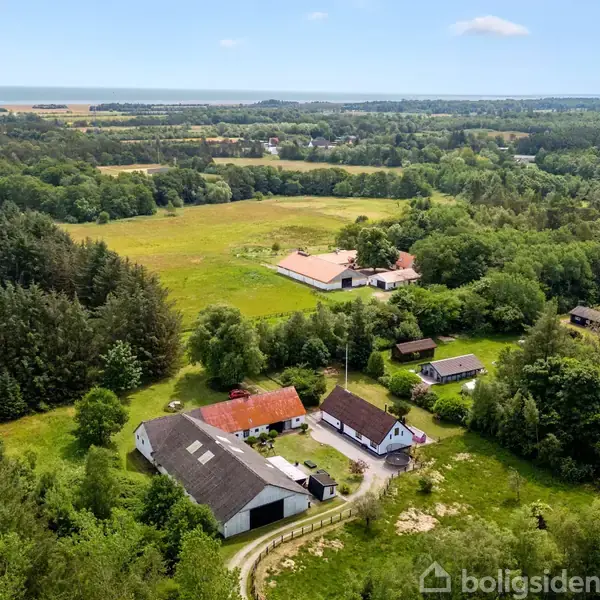Gårde ligger spredt på en frodig, grøn ejendom med flere bygninger omgivet af tæt skov og åbne marker, beliggende i et landligt område med en langstrakt horisont.