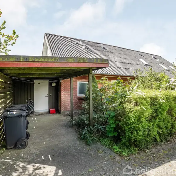 Carport foran et enfamiliehus med røde mursten. To skraldespande står ved indgangen. Hække og træer omkranser området. Dør mærket med nummer 14. Himmel let overskyet.