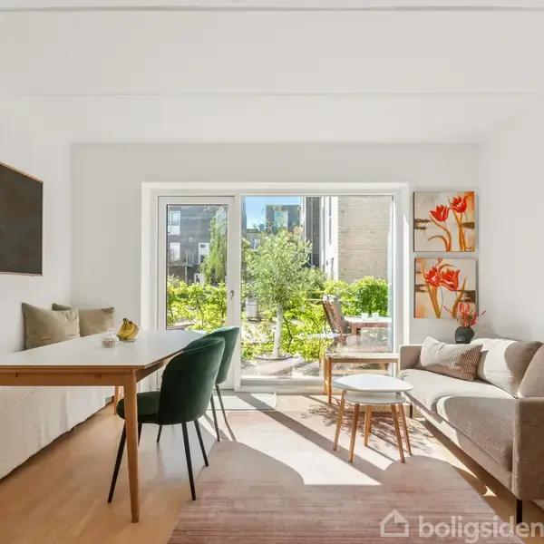 Stue med en beige sofa til højre, et langt spisebord med to stole til venstre, og store glasdøre i baggrunden, der åbner ud til en grøn terrasse.