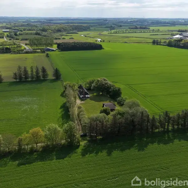 Brændeovn opvarmer hyggelig spisestue med rundt bord og stole i rustik indretning. Køkken ses i baggrunden med moderne elementer. Vinduer lukker dagslys ind og udsigt til naturen.