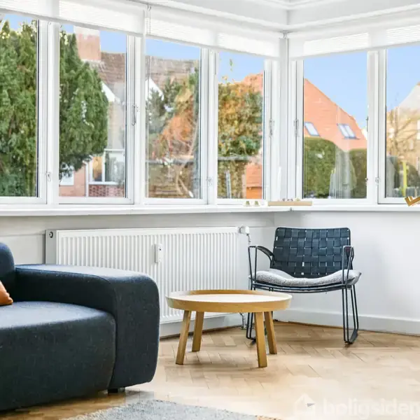 Sort sofa står roligt ved siden af et rundt træbord og en metalstol i en lys stue med store vinduer. Udenfor ses huse og træer.