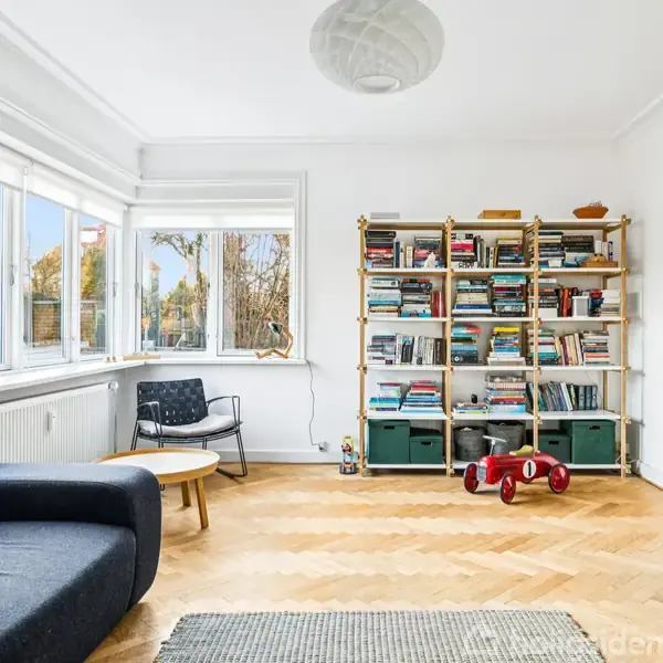 En lys stue med træhylde fyldt med bøger ved væggen, blå sofa med orange pude, rundt træbord og stor vinduesrække. Gulvet har sildebensparket, og en lille rød legetøjsvogn står på gulvet.