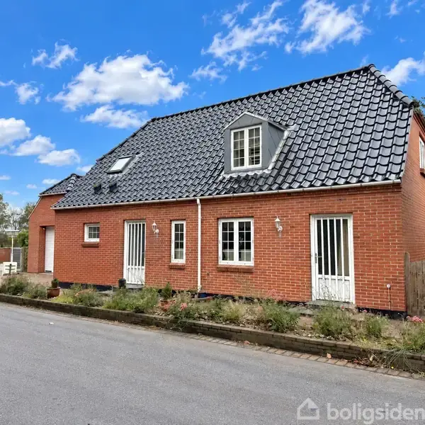 Rødt murstenshus med sort tegltag står foran en vej. Huset har flere døre og vinduer. Blå himmel og træer ses i baggrunden.