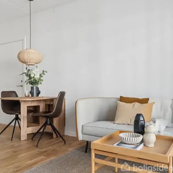 Sofa med puder står ved et sofabord med dekoration, mens et spisebord med stole og en lampe ses i baggrunden. Trægulv og hvide vægge skaber en lys atmosfære.