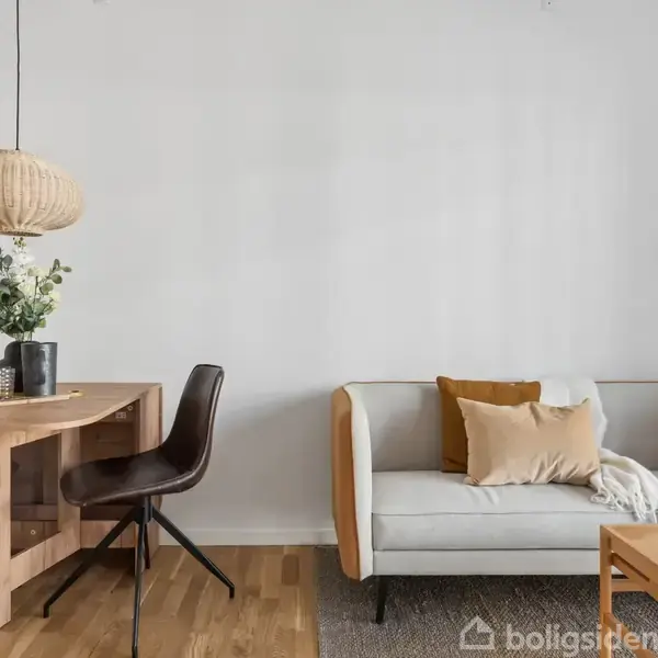En stue med en lys sofa, der har puder og et tæppe, står tæt på et træbord med stole. En pendellampe hænger over bordet, omgivet af en neutral indretning.