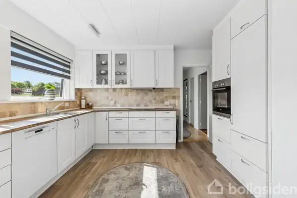 Et lyst køkken med hvide skabe og skuffer, tre indbyggede ovne samt en vask under et stort vindue. Der ligger et rundt tæppe på trægulvet.