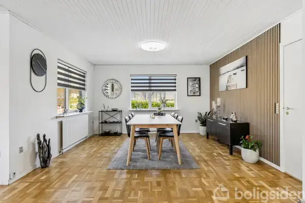 Spisebord med fire stole står på et gulvtæppe i lyse spisestue med trægulv og hvide vægge. To store vinduer har stribede rullegardiner, og der hænger ure og billeder på væggene.