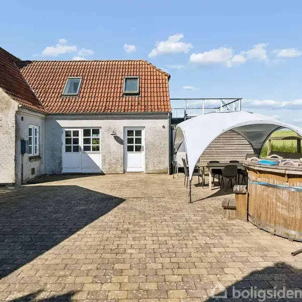 Et gårdspladsområde med brosten, hvor en hvid bygning med rødt tegltag står til venstre. Til højre er der et udendørs badekar omgivet af en baldakin og havemøbler under åben himmel.