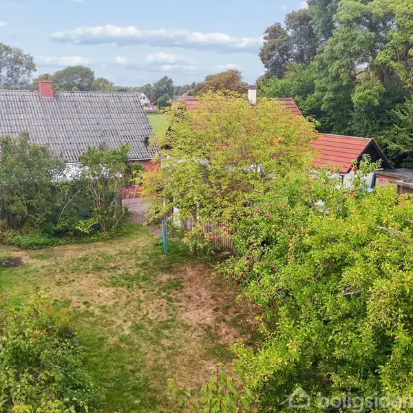 To huse med røde tagsten står blandt frodig vegetation i en haveomgivelse. Omkringliggende træer og buske danner en grøn ramme. Det er en fredelig og landlig scene.