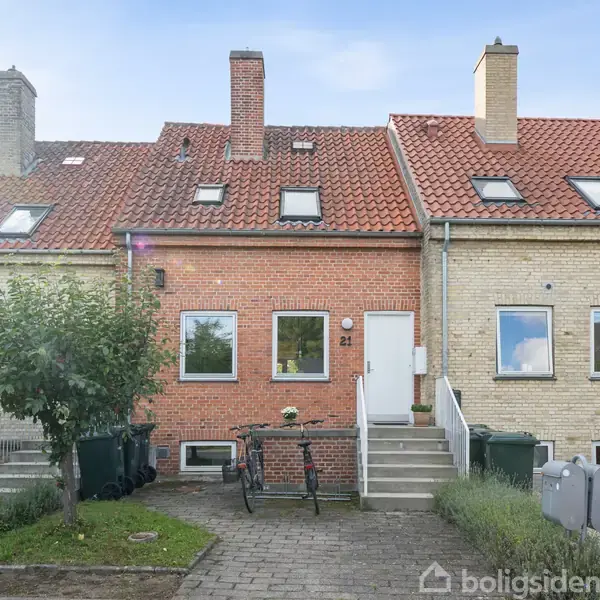 Rækkehus i røde og gule mursten, med rødt tegltag. Cykler står foran nummer 21. Huset har to vinduer og en trappe op til døren, omgivet af en have.