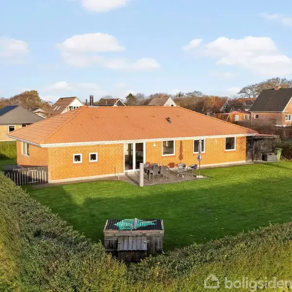 Et rødt murstenshus med rødt tag står i en velplejet grøn have omgivet af hække. Huset har en terrasse med siddemøbler. Nabolaget består af lignende huse.
