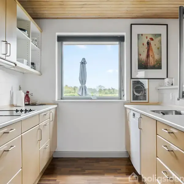 Et lyst køkken med flisebeklædte skabsfronter, integrerede ovne og keramisk kogeplade; en vask ved højre bordplade. Et vindue med udsigt til haven og et paraplyformet objekt udenfor.