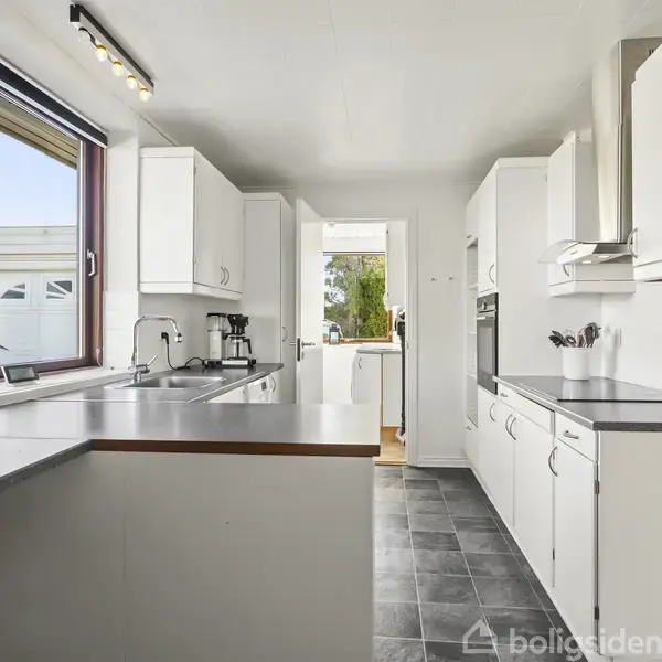 Et moderne køkken med hvide skabe og grå bordplader i en U-form. Der er en stor vinduesåbning til venstre, og et sort gulvfliser. Adgang til næste rum i baggrunden.