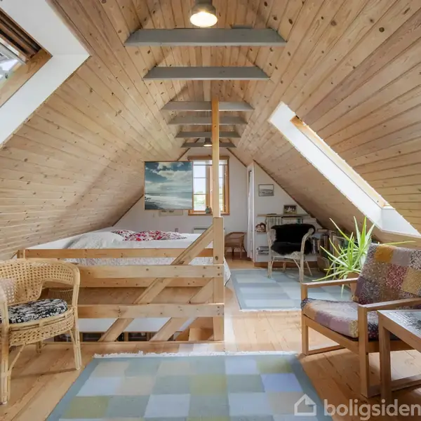 Skråloft-værelse med trægulv og trævægge. En seng står til venstre, mens en stol og et bord med blomster er til højre. Store ovenlysvinduer giver naturligt lys.