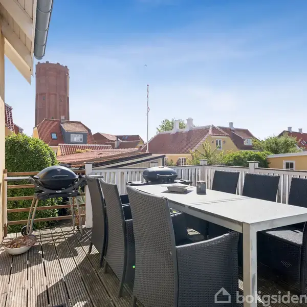 En terrasse med et spisebord omgivet af sorte stole og en grill. Solskin og udsigt over hustage og en rødstensbygning i en bymæssig omgivelser.