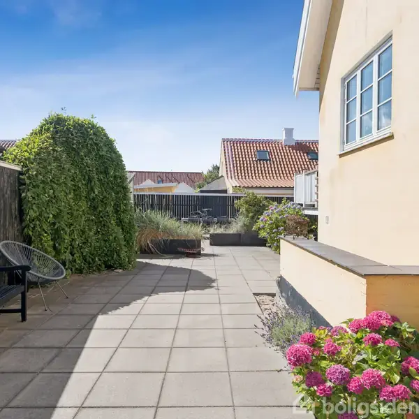 Bænke og stole står på en flisebelagt terrasse ved siden af en gul husvæg. Rosa blomster og grønne buske tilføjer farve, mens omkringliggende bygninger ses i baggrunden under en blå himmel.