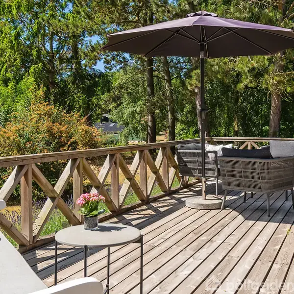 Et terrasseområde med træmøbler og en stor parasol står på et trædæk. Bordet har en blomsterpotte. Omgivet af grønne træer og buske, skaber en hyggelig udendørs atmosfære.