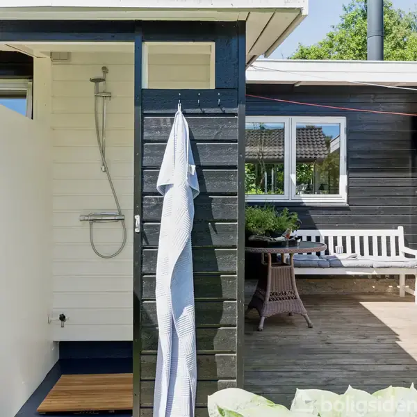 Udendørs bruser med et ophængt håndklæde ved siden af et sort træsommerhus. Bruseren er placeret på en træterrasse med en bænk og bord i baggrunden under en klar blå himmel.