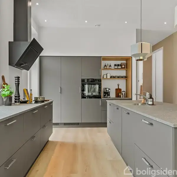 Et moderne køkken med mørkegrå skabe og skuffer. Køkkenøen har en lys bordplade. Ovn og hylder med glasflasker findes ved væggen. Store vinduer giver naturligt lys.