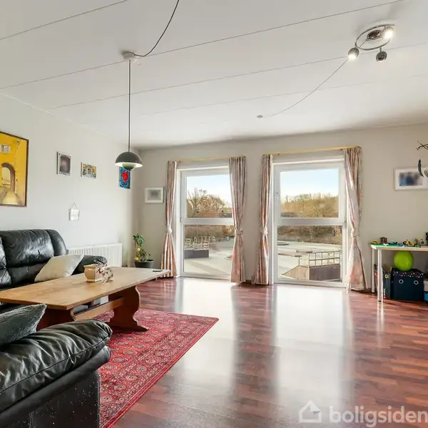Sort sofa står ved et træbord på et rødt tæppe i en stue med træparketgulv. To store vinduer slipper lys ind; legetøj er arrangeret på hylden til højre.