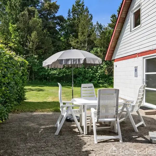 En udendørs terrasse med et hvidt plastbord og fire stole står under en parasol. Terrassen tilhører et hus med hvid facade og rød kant. Græsplæne og træer omkranser området.