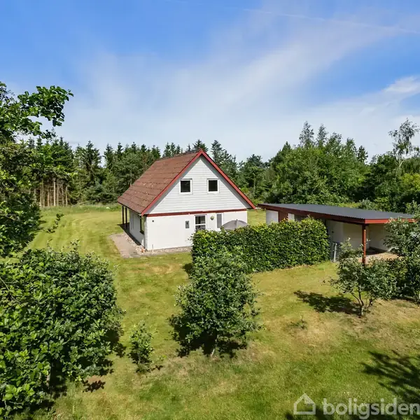 Et hvidt hus med rødt tag står i en grøn have omgivet af træer og buske, under en blå himmel. Ved siden af huset er en carport tilknyttet.