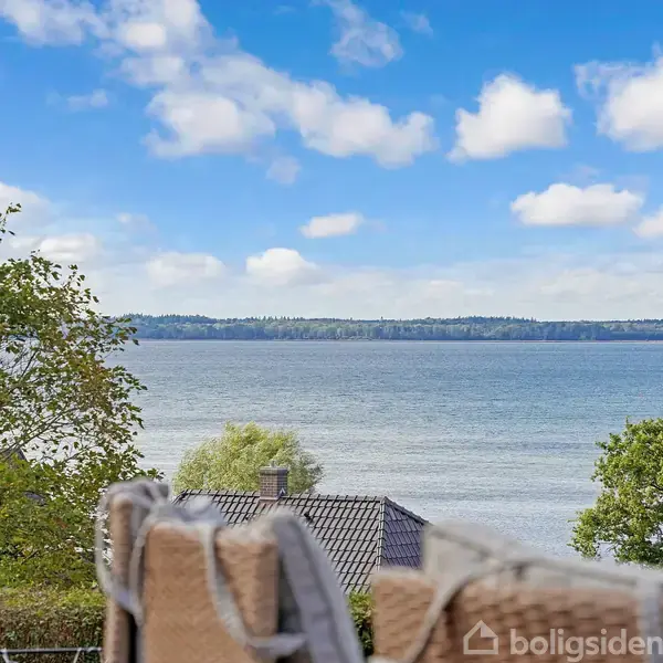 En flettet kurv står på en altan med udsigt over vandet, omkranset af træer. Lyseblå himmel med lette skyer danner baggrund.