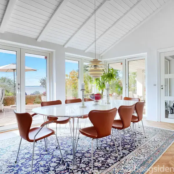 Et moderne spisebord omgivet af seks brune stole står på et blåt orientalsk tæppe i en lys spisestue med store vinduer, der åbner ud mod en terrasse med udsigt.