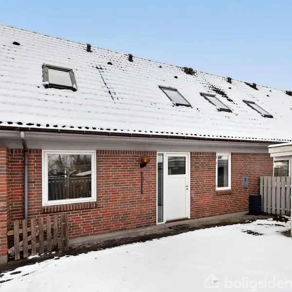 Et rødt murstenshus med et snedækket tag og flere ovenlysvinduer. Husets hoveddør er hvid, omgivet af sne i en indkørsel. Hegn og carport ses i baggrunden.