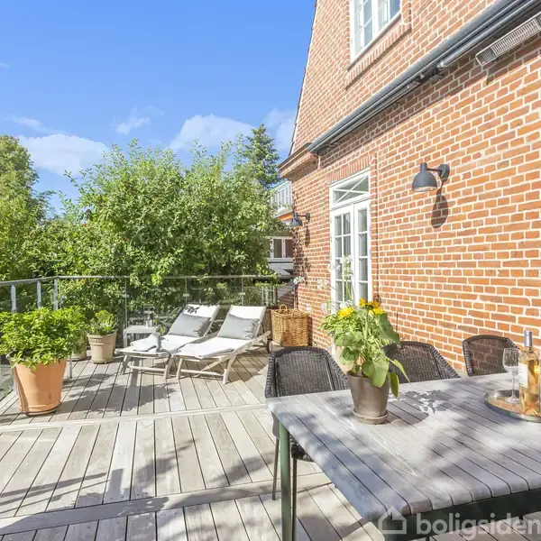 Terrasse med havebord og stole, står på trædæk ved rødstenshus. To liggestole er placeret ved siden af store grønne planter. Blå himmel og træer omkranser området.