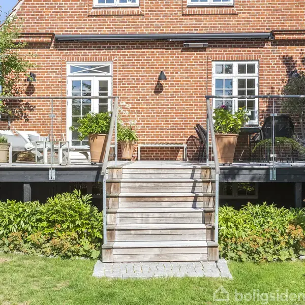 Rødt murstenshus med en terrasse i træ, hvor blomsterkrukker står ved glasrækværk. Trappen fører ned til en frodig have. Store vinduer reflekterer sollys.
