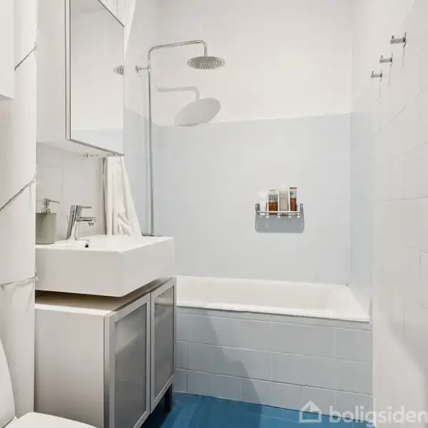 Badeværelse med toilet, håndvask og spejlskab. Badekar under bruser. Hvide flisevægge og blå gulv. Vægophængt hylde over badekarret med shampoo og sæber.