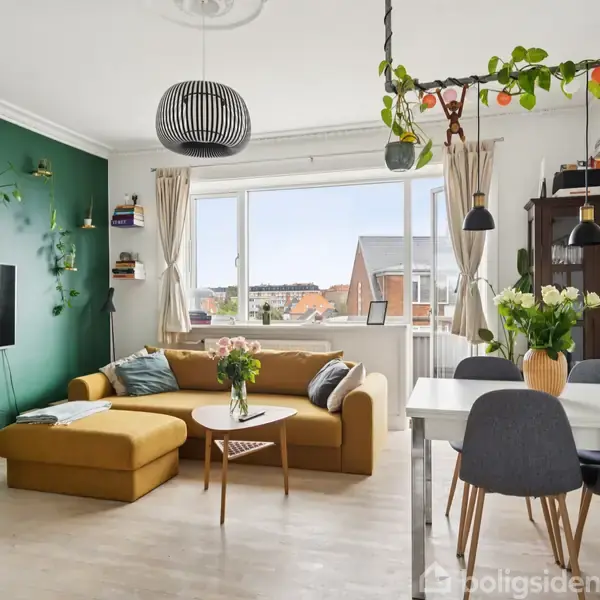 En stue med en gul sofa står ved en grøn væg med et tv. Et spisebord med sorte stole er placeret under planter, og store vinduer viser en udsigt over naboens tage.