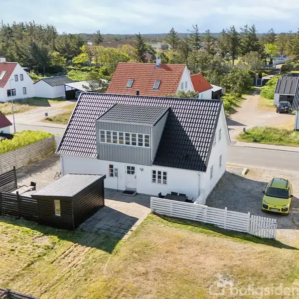 Huset, en hvid bygning med sort tag og tilhørende skur, står i en græsplæne omkranset af hvidt stakit. En gul bil er parkeret foran på en stille vej.