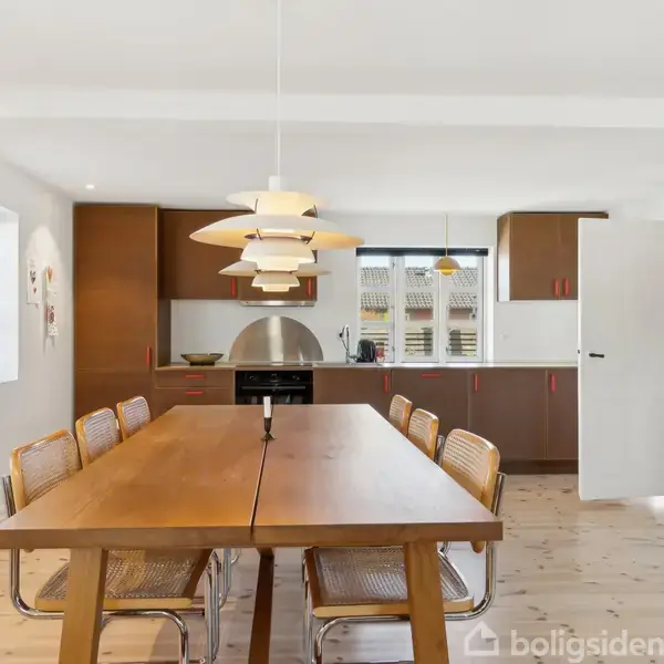 En lang træspisebord står centralt med seks fletstole omkring. En moderne lampe hænger ovenover. Bagved er et åbent køkken med hvid bordplade og træskabe. Vinduer lader lys strømme ind.