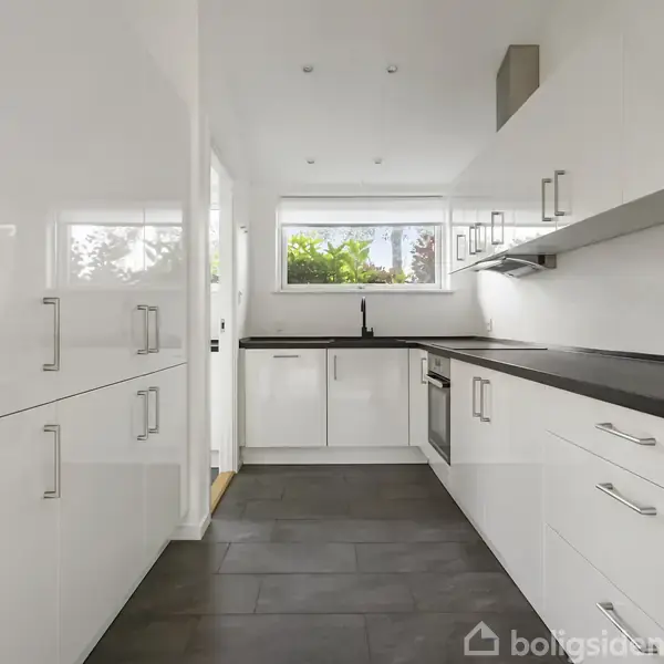 Et moderne køkken har hvide skabe og sorte bordplader. Et vindue i baggrunden giver udsigt til grønne planter. Rummet er lyst og minimalistisk indrettet.