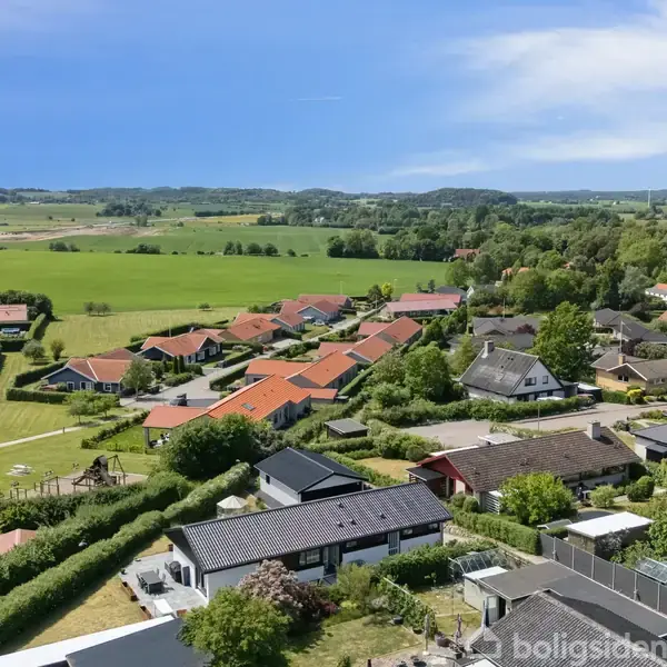 Huse med røde tage ligger spredt blandt grønne træer i et landligt boligområde. I baggrunden ses marker og skov under en blå himmel.