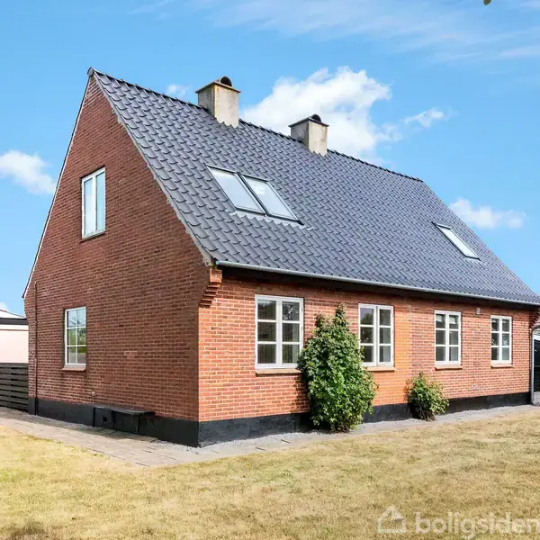 Et rødt murstenshus med sort tag står i en velplejet have. Huset har flere vinduer og to skorstene. Himmelen er blå med enkelte skyer.
