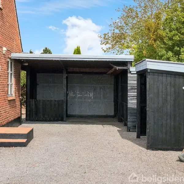 Carport står tom ved siden af rødt murstenshus, med sortmalet skur til højre. Grusbelagt gårdsplads med træer i baggrunden.