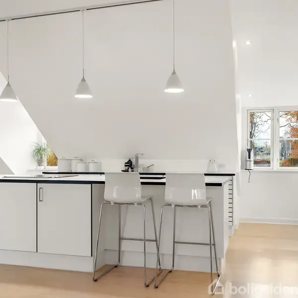 Et moderne, hvidt køkken med en køkkenø og to barstole, oplyst af tre loftslamper. I baggrunden ses et vindue med udsigt til træer med efterårsblade.