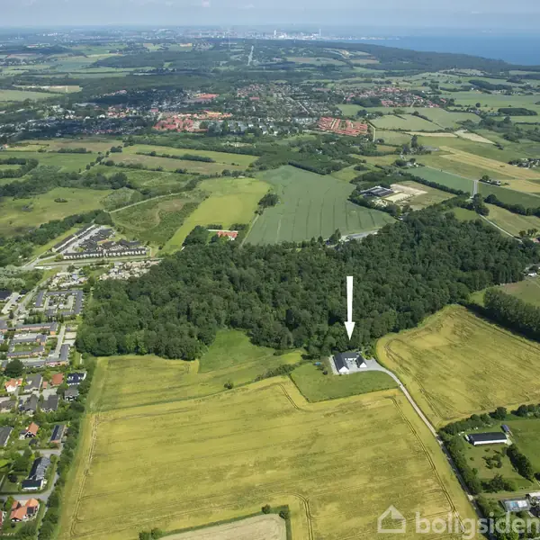 Hus vist fra oven, markeret med en pil, omgivet af grønne marker og skov. I baggrunden ses en mindre by og kystlinjen med havudsigt under en klar himmel.